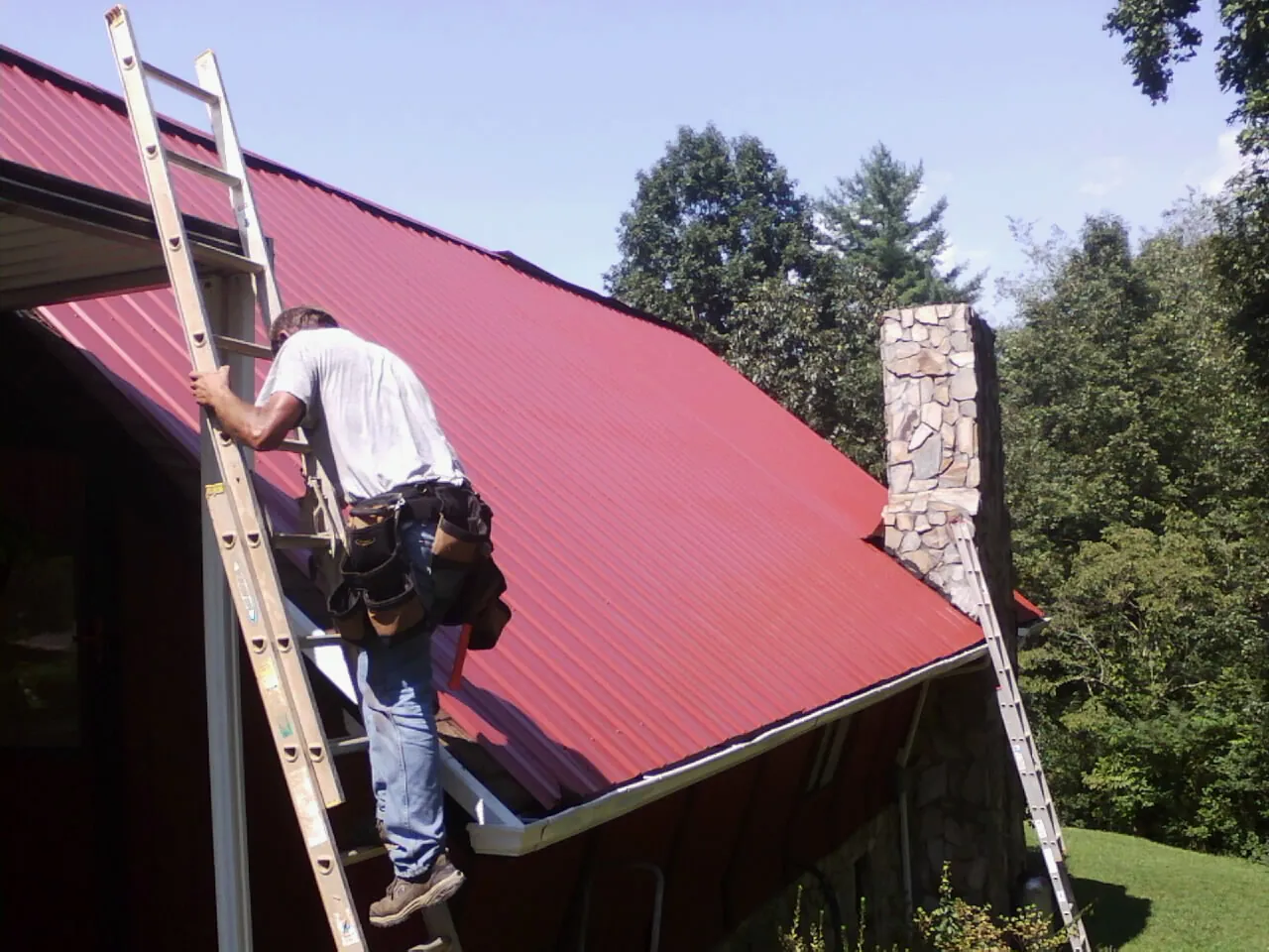 Roofer on a ladder beside a red metal roof and stone chimney — Soffit & Fascia Repair by Roofing Brothers in Jamesport